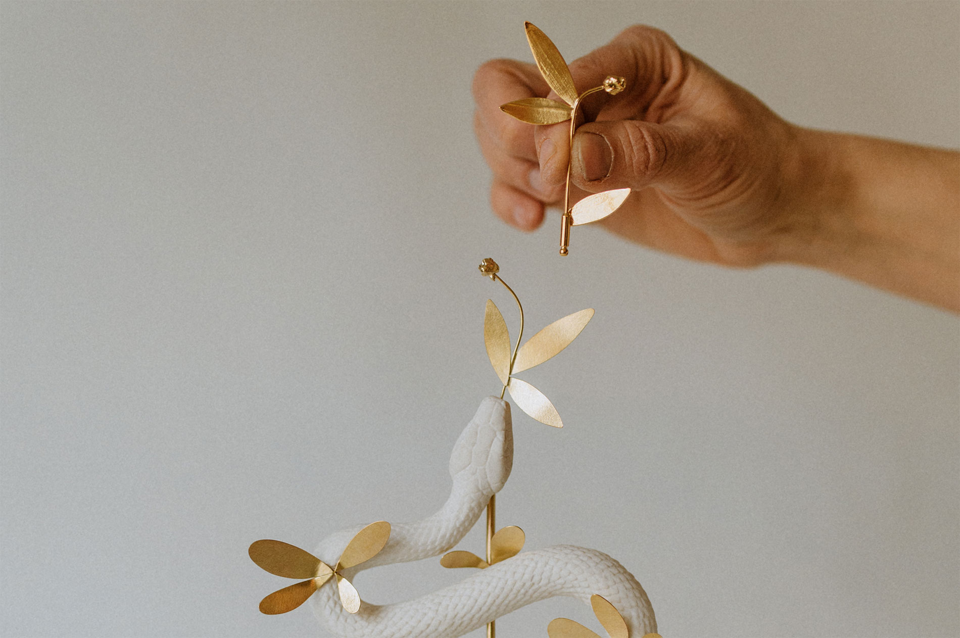 Main posant une broche feuillage en laiton doré dans la tête d'un serpent en porcelaine, sculpture Chimeria, Les Aubépines